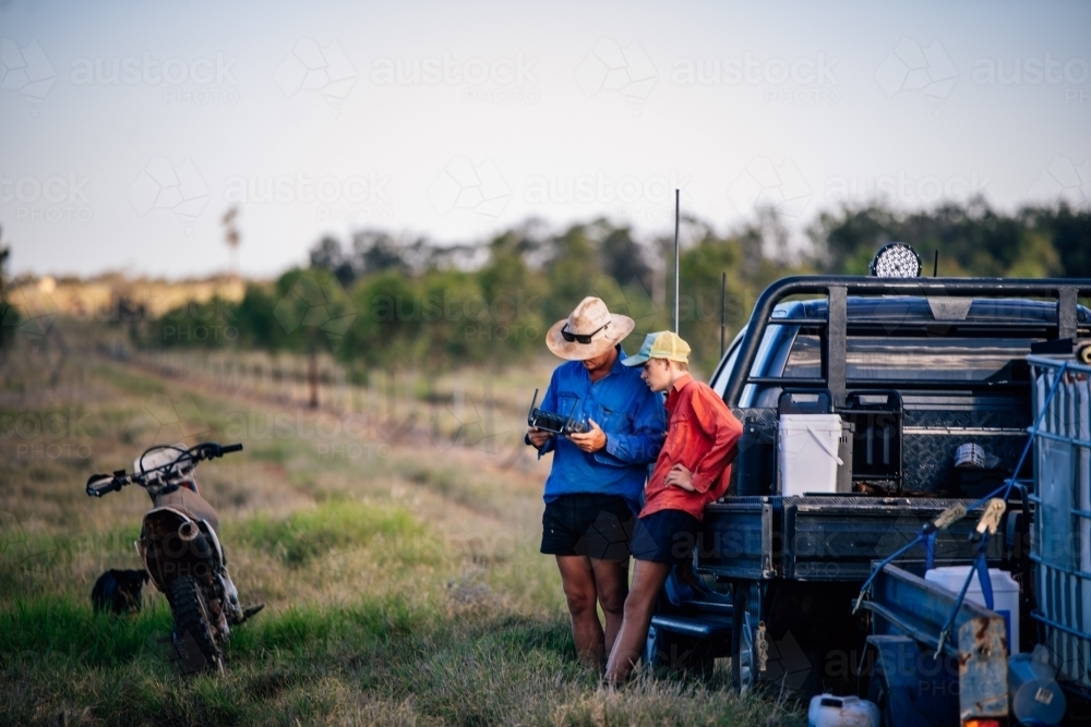 2 workers looking at a device in the paddock with a work ute and 2 wheel motorbike - Australian Stock Image