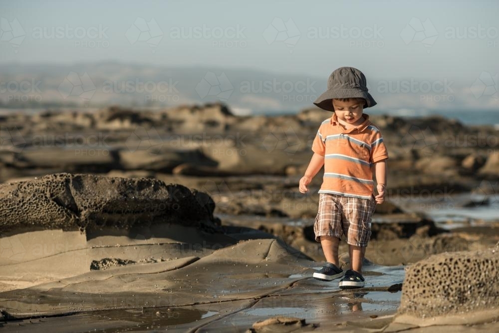 2 year old mixed race boy looks for shells on a rocky coast - Australian Stock Image