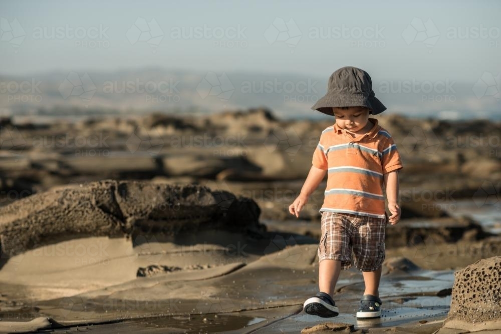 2 year old mixed race boy looks for shells on a rocky coast - Australian Stock Image