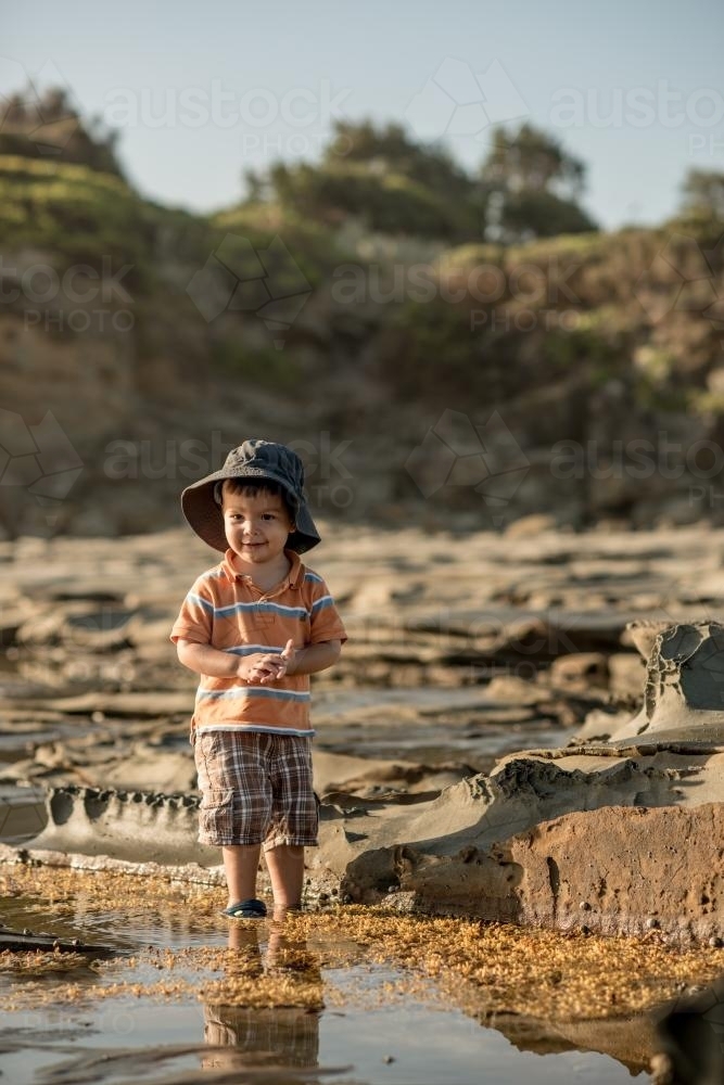 2 year old mixed race boy looks for shells on a rocky coast - Australian Stock Image