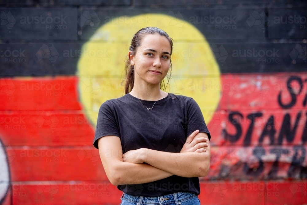 20-something Aboriginal woman standing with her arms crossed in front of a painted flag - Australian Stock Image