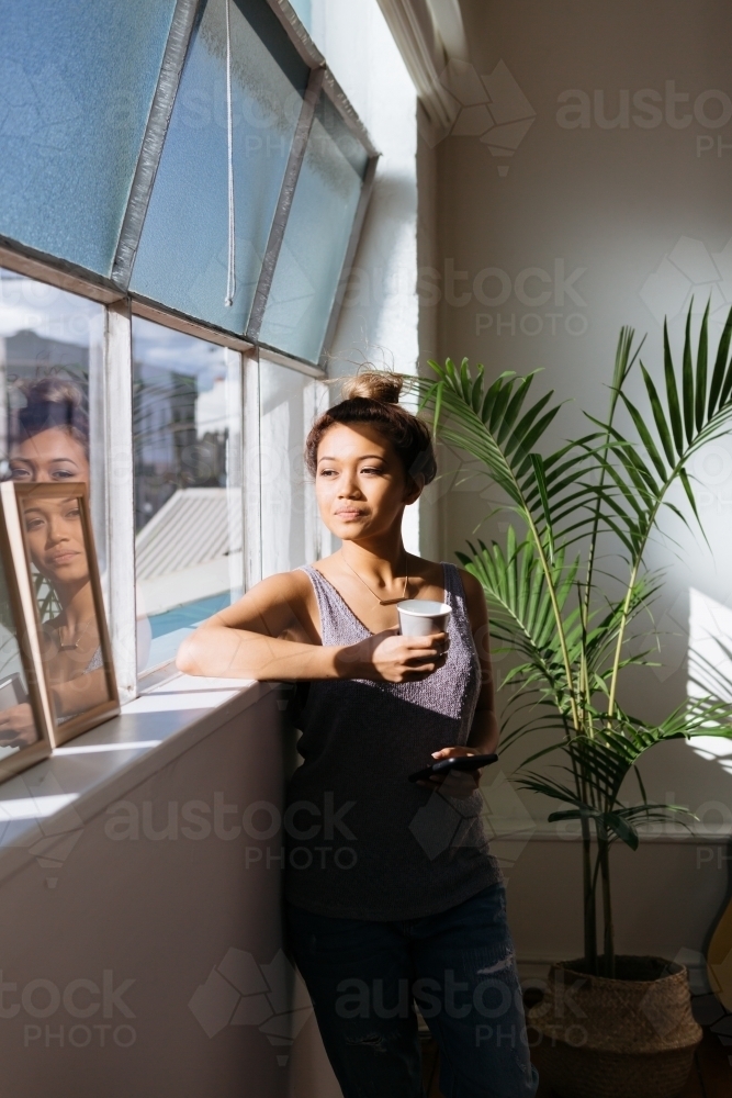 20 something woman with a coffee looking out her window in contemplation - Australian Stock Image