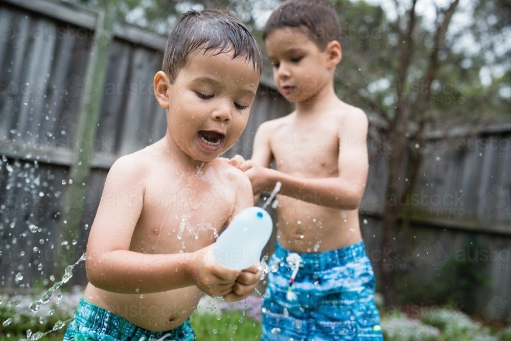 3 year old and 6 year old mixed race brothers play excitedly with water bombs in suburban backyard - Australian Stock Image