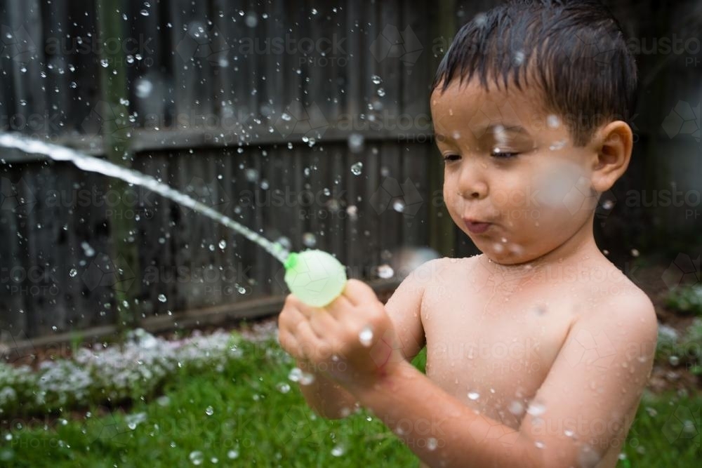3 year old mixed race boy plays excitedly with water bombs in suburban backyard - Australian Stock Image
