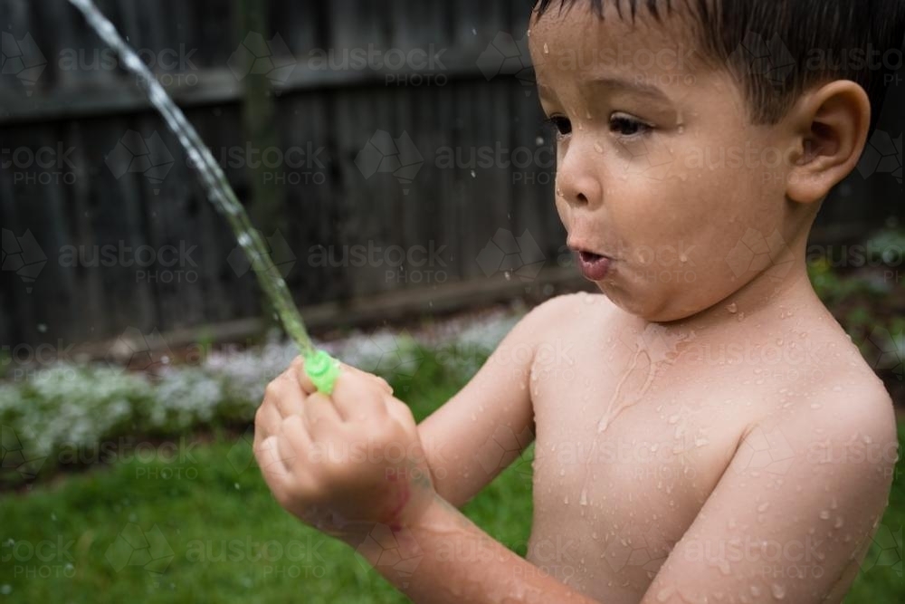 3 year old mixed race boy plays excitedly with water bombs in suburban backyard - Australian Stock Image