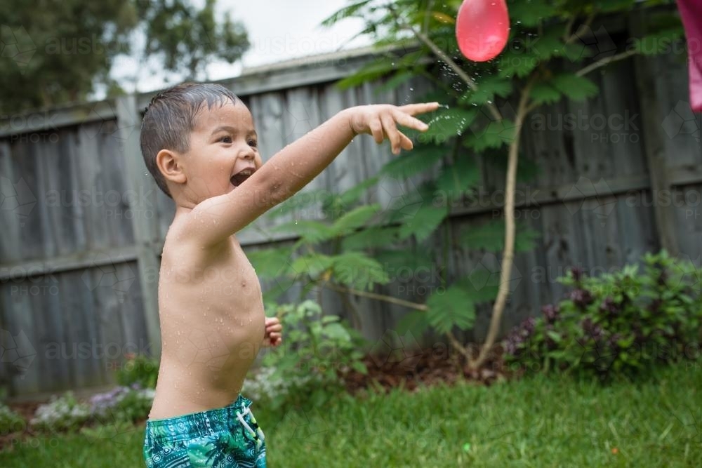 3 year old mixed race boy plays excitedly with water bombs in suburban backyard - Australian Stock Image