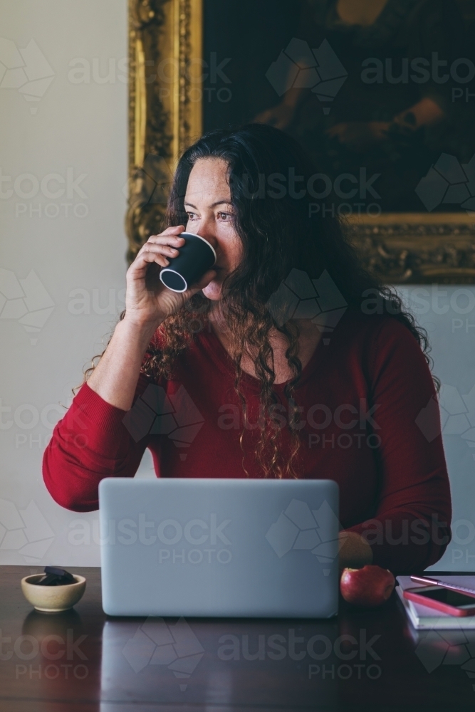 40 something woman working from home on laptop, drinking coffee - Australian Stock Image
