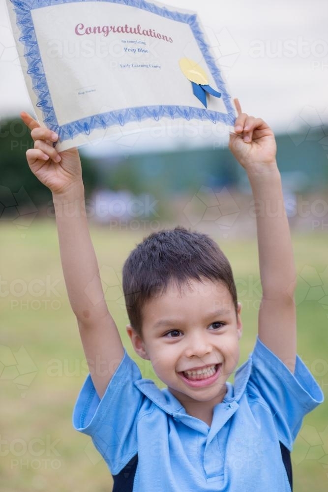 5 year old boy celebrates his graduation from Prep by holding his certificate above his head - Australian Stock Image