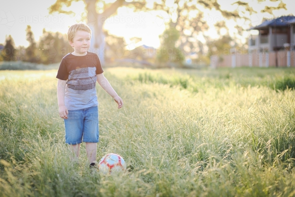 6 year old boy playing soccer in long grass at the park. Outdoor fun in nature, - Australian Stock Image