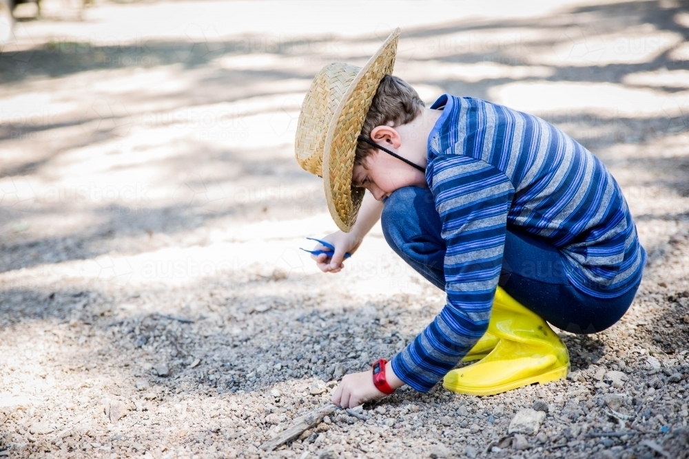 Child in gumboots and hat holding tweezers looking through rocks and dirt on ground - Australian Stock Image