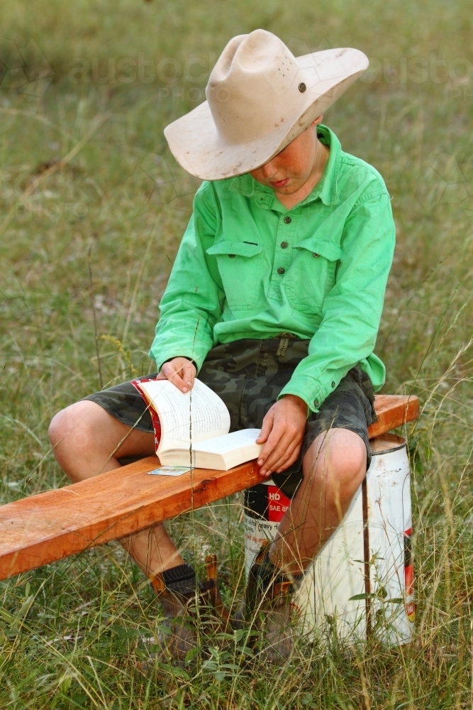 A 10 year old country boy reading a book on a makeshift bench in a paddock - Australian Stock Image