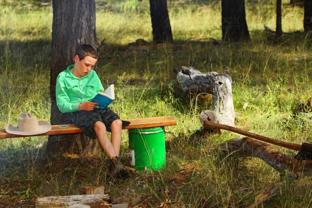 A 10 year old country boy reading a book on a makeshift bench next to a fire among a stand of trees - Australian Stock Image