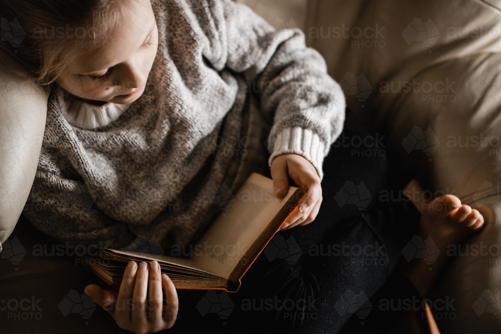 A blonde caucasian girl sitting curled up on a chair reading a book - Australian Stock Image