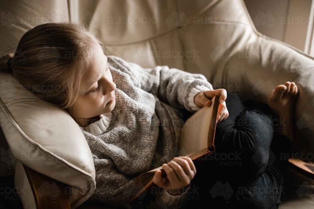 A blonde caucasian girl sitting curled up on a chair reading a book - Australian Stock Image