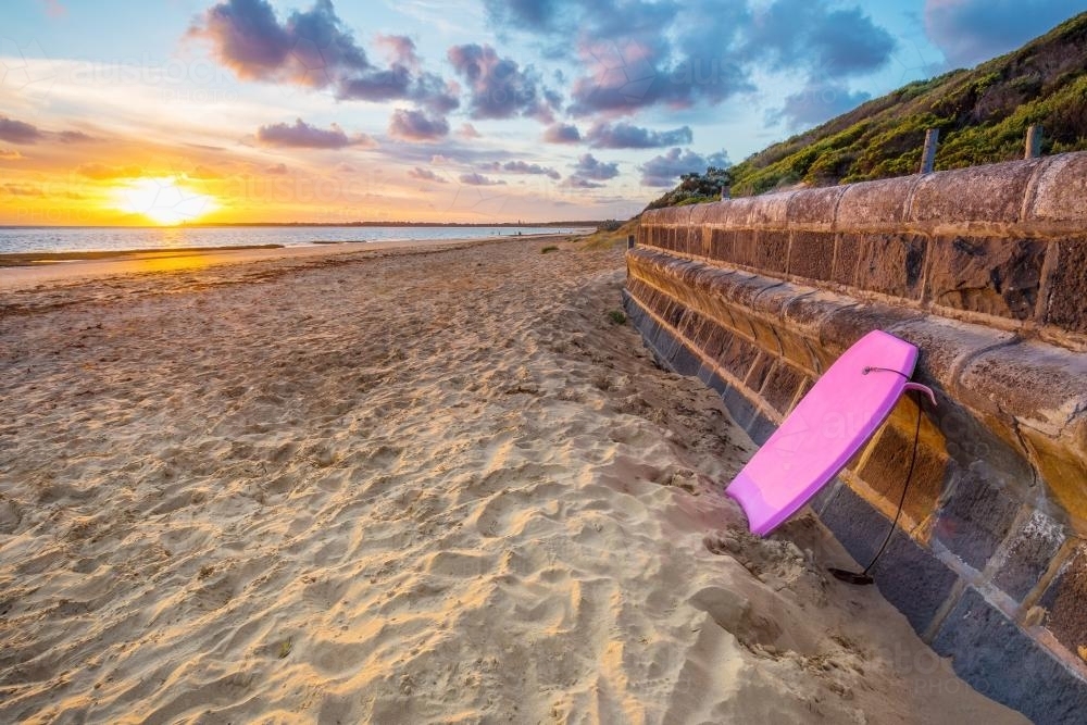A boogie board is left leaning against a beach wall at sunset - Australian Stock Image