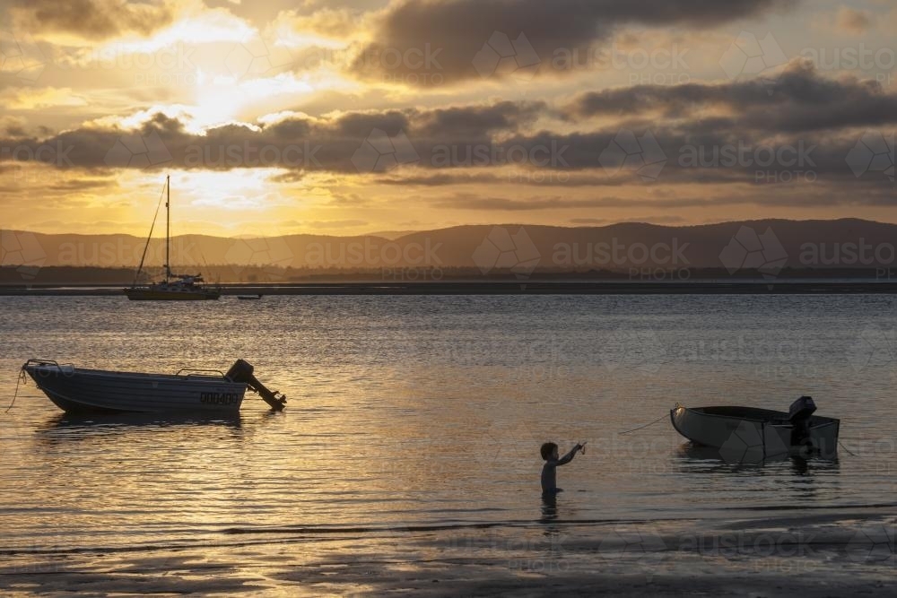 A boy and some boats in the water at sunset. - Australian Stock Image