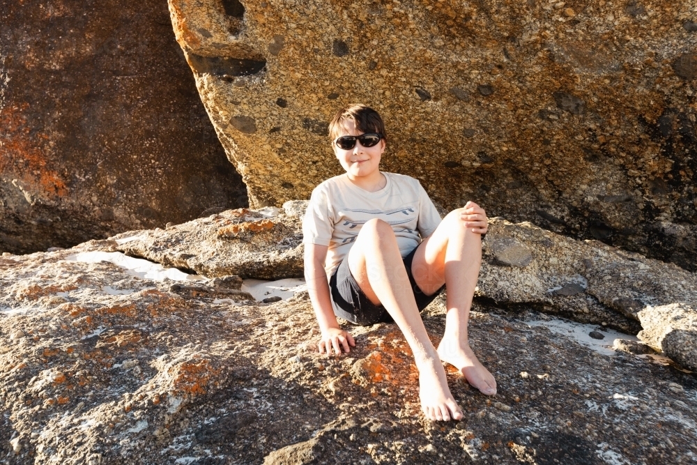a boy child wearing sun glasses sitting on boulder rocks at Squeaky beach in Wilson's Promontory Sou - Australian Stock Image