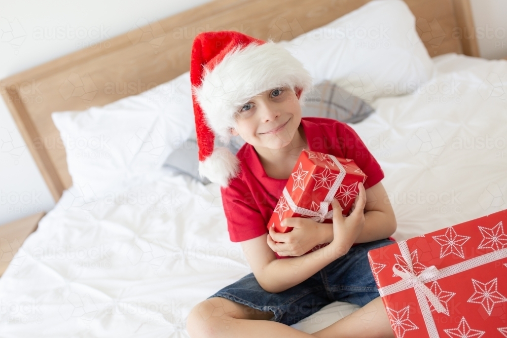 A boy sitting on a bed wearing a red shirt and a Santa hat at christmas time holding his presents - Australian Stock Image