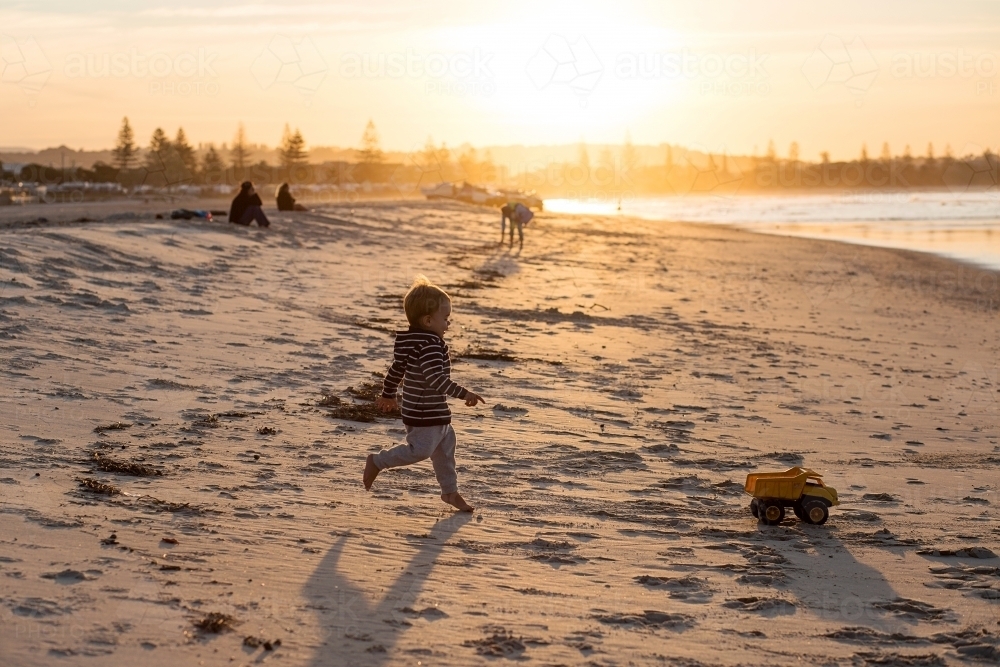 A Boy toddler, running after a toy truck on the beach at sunset - Australian Stock Image