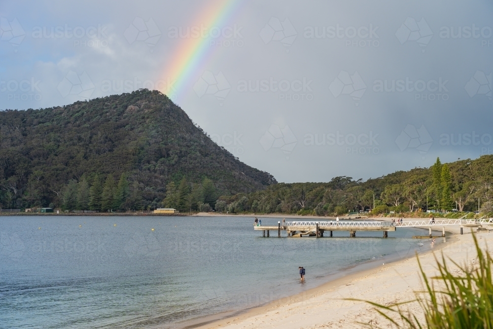 A bright rainbow over a coastal mountain behind a curved sandy beach with a jetty - Australian Stock Image