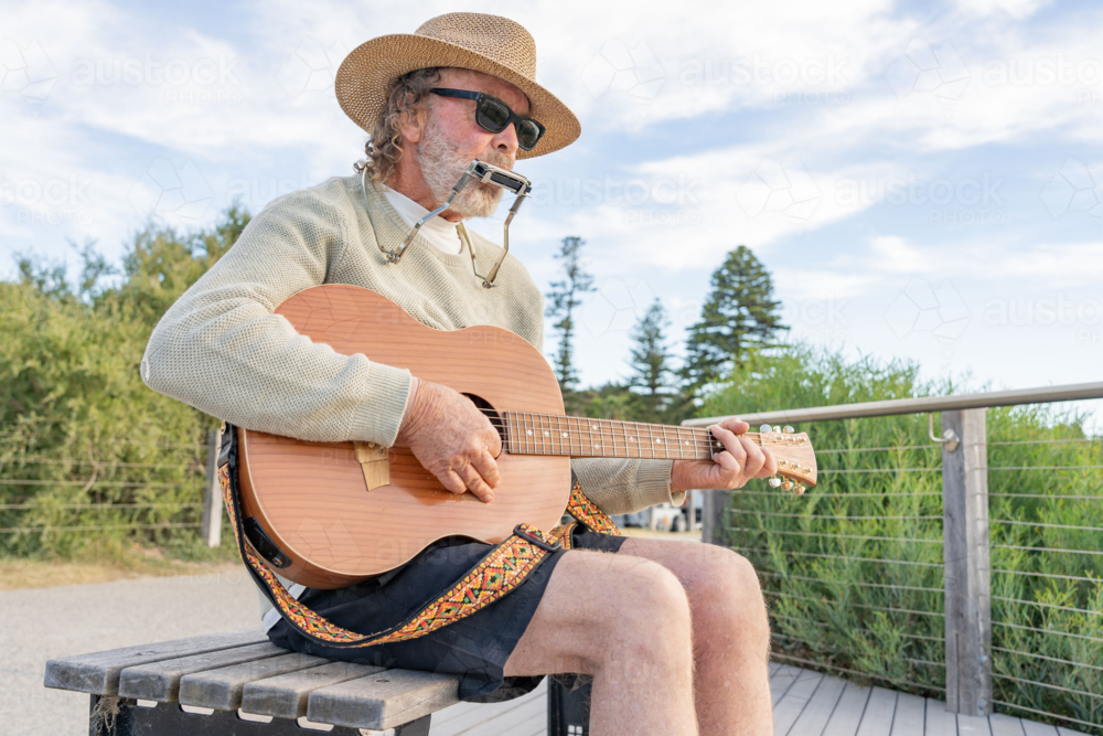 A busker wearing a hat playing a guitar and harmonica on a park bench - Australian Stock Image
