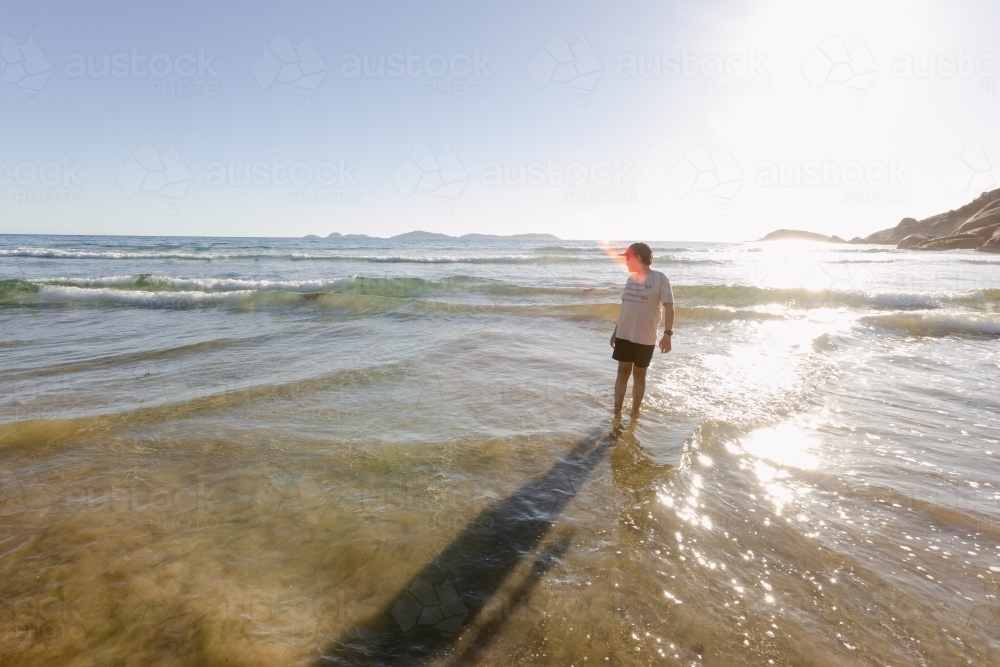 A Caucasian boy child wearing a hat standing alone in the water at an australian beach with waves - Australian Stock Image