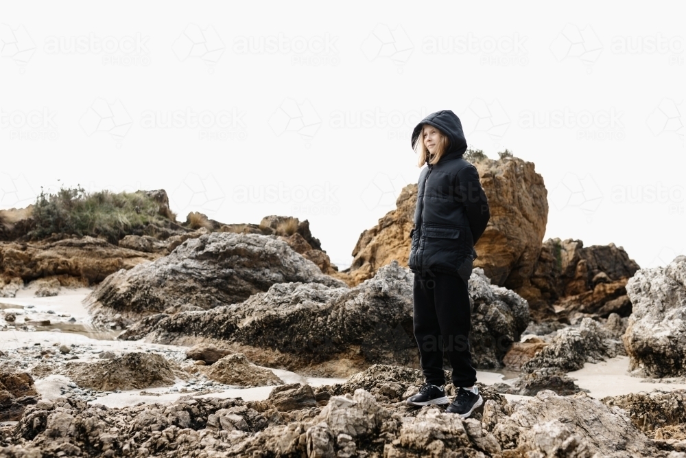 A causcasian blonde girl wearing a warm jacket standing on rocks at a beach in winter - Australian Stock Image
