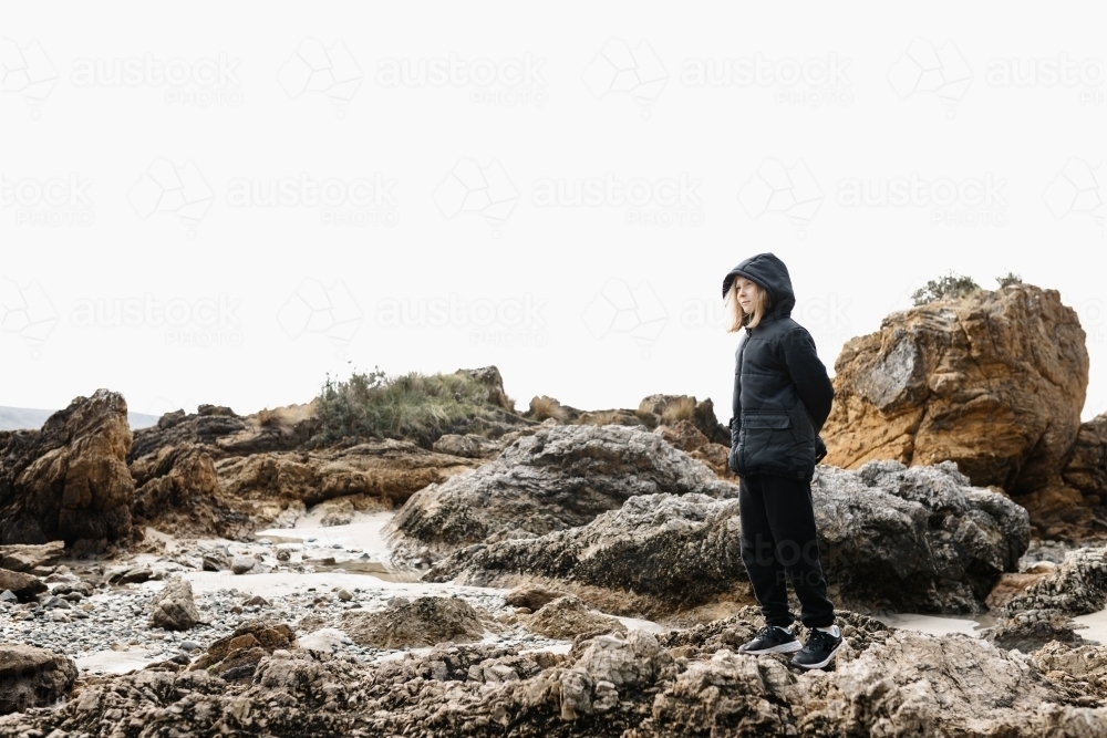 A causcasian blonde girl wearing a warm jacket standing on rocks at a beach in winter - Australian Stock Image