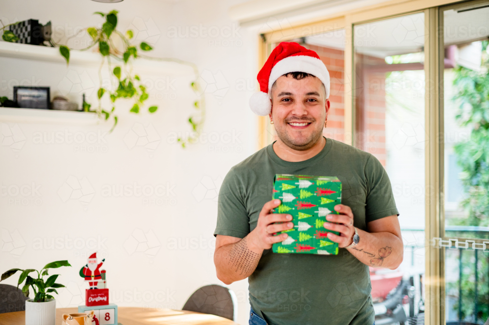 A cheerful man in a Santa hat smiles while holding a wrapped present in a festive living room - Australian Stock Image