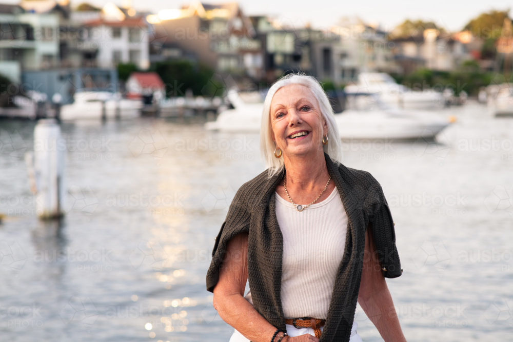 A cheerful senior woman with white hair stands by a serene waterfront, smiling warmly : Austockphoto A cheerful senior woman with white hair stands by a serene waterfront, smiling warmly - Australian Stock Image