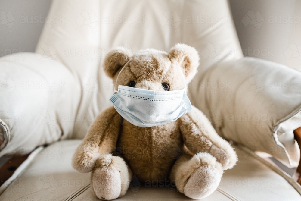 A child's teddy bear sitting on a chair wearing a facemask - Australian Stock Image