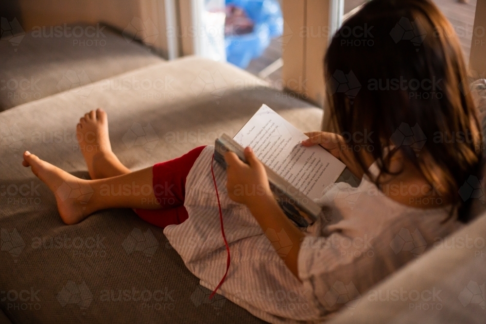 A child sitting on a couch in the living room reading a bedtime storybook - Australian Stock Image
