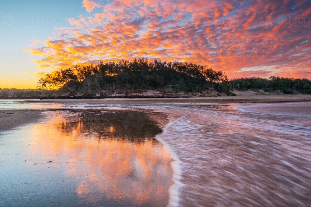 A colorful coastal sunset reflected on a wet sandy beach with waves rushing over it - Australian Stock Image