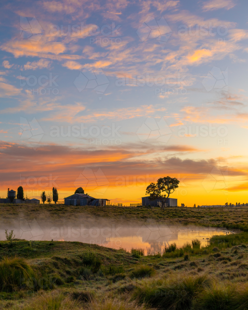 A colourful sunrise with the silhouette of two old sheds and trees in dam on farm - Australian Stock Image