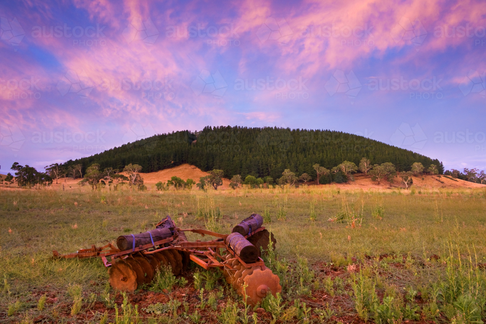 A colourful sunset sky over a rusty plough in a paddock in front of a tree covered mountain - Australian Stock Image