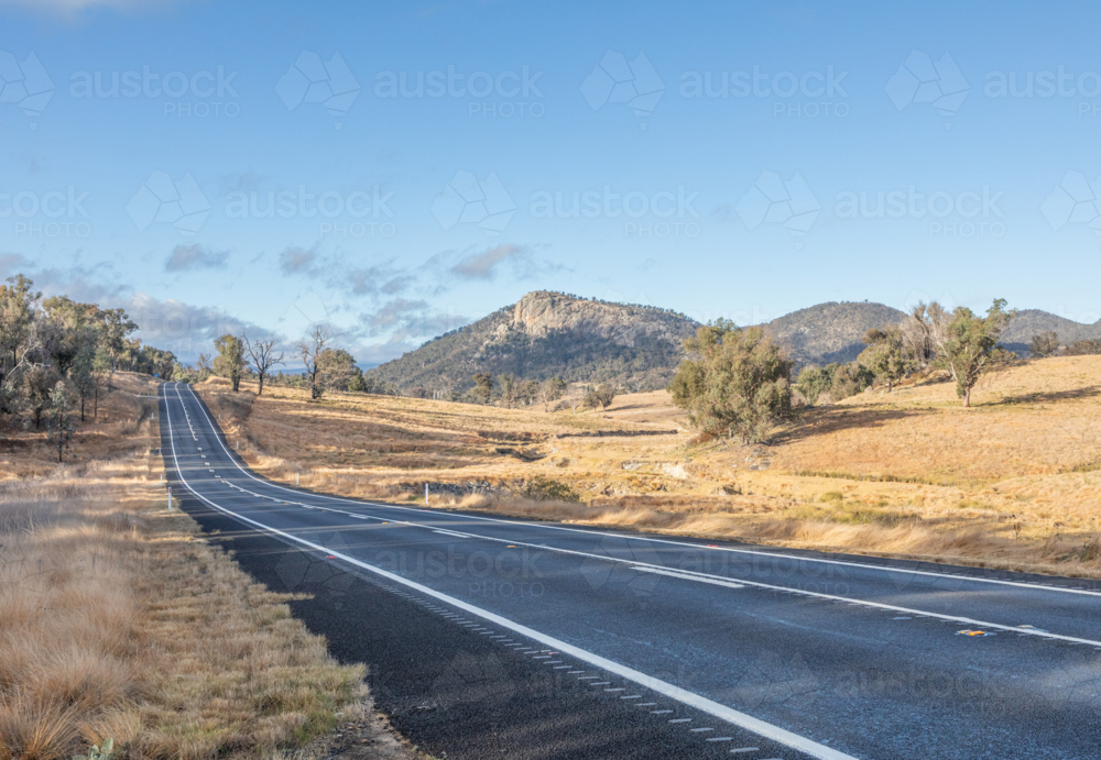A country highway with a granite rock in the distance underneath a blue sky - Australian Stock Image