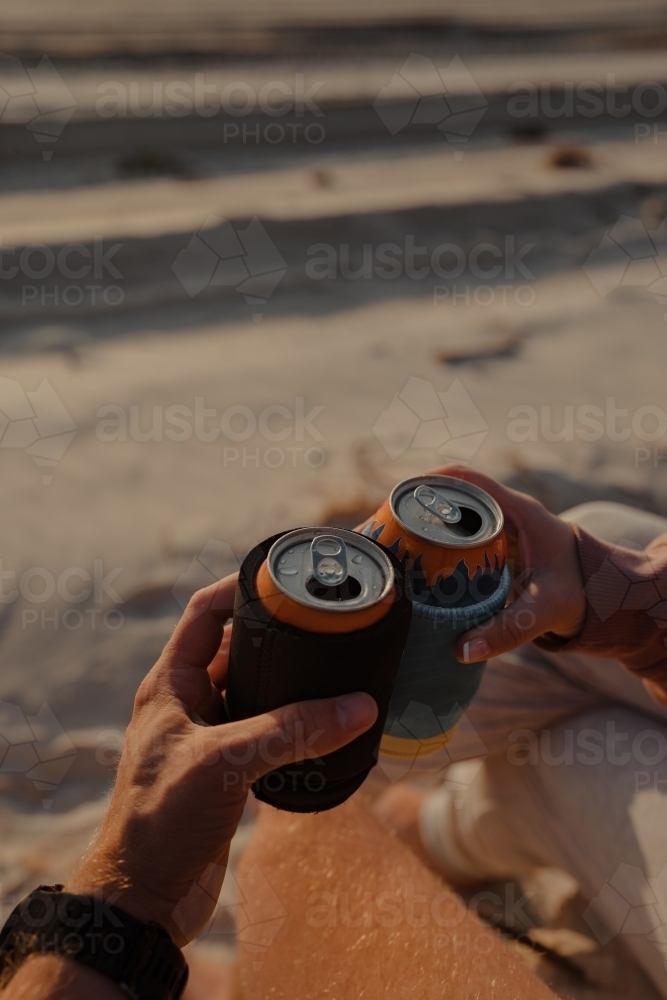 A couple cheersing two cans of drink at sunset on the beach. - Australian Stock Image