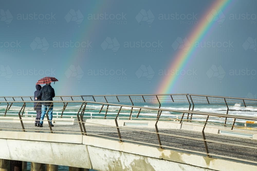 A couple walk on a jetty with an umbrella looking at a rainbow - Australian Stock Image