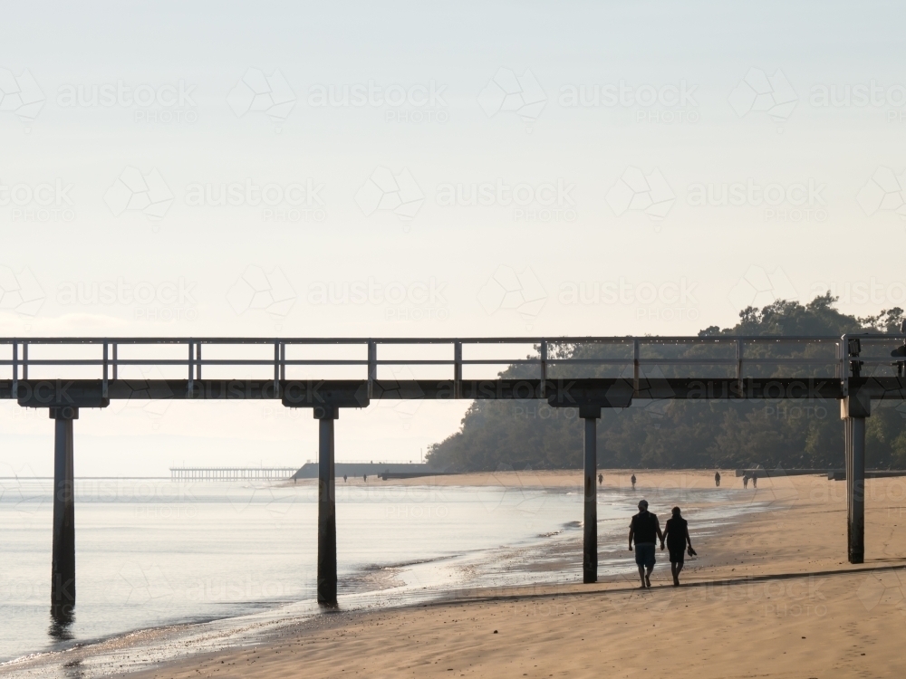 A couple walking hand-in-hand on the beach under a jetty - Australian Stock Image