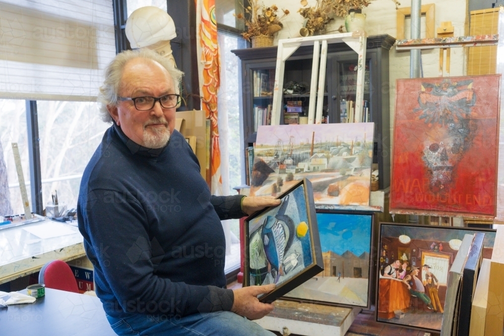 A distinguished male artist sitting holding a painting in his studio - Australian Stock Image