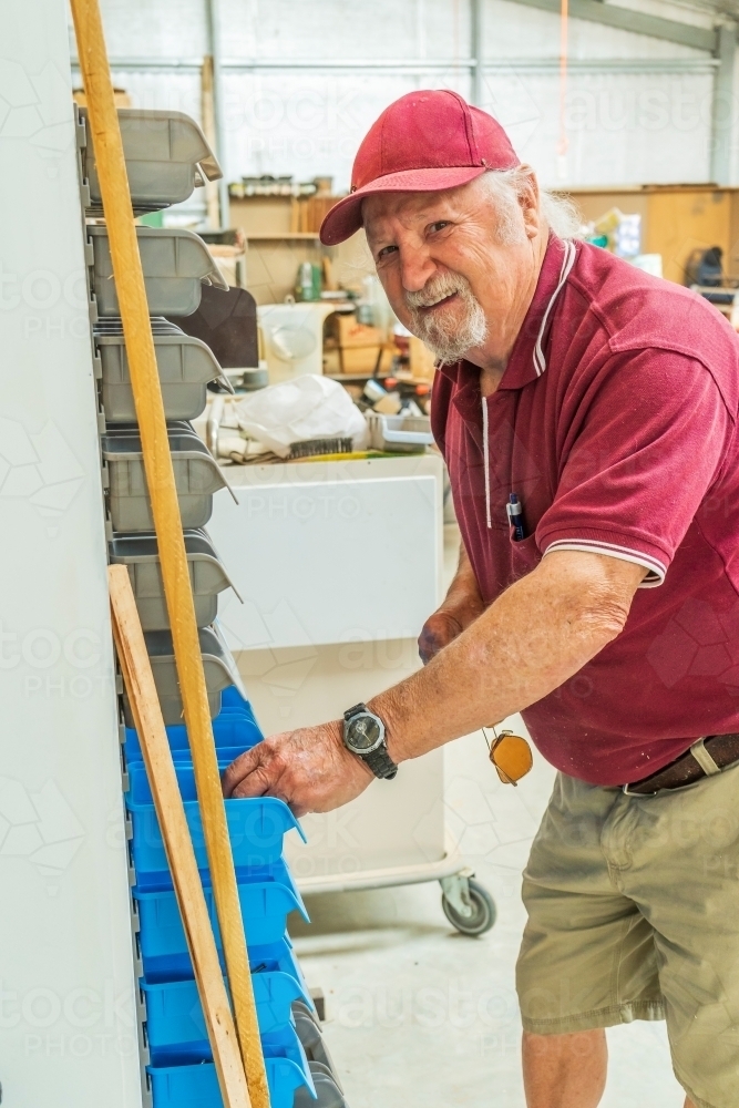 A elderly bearded handyman smiling while searching through racks on a wall in a Men's shed - Australian Stock Image