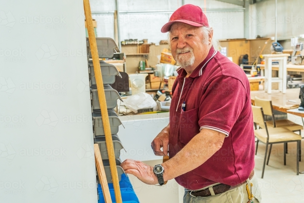 A elderly bearded handyman smiling while searching through racks on a wall in a Men's shed - Australian Stock Image