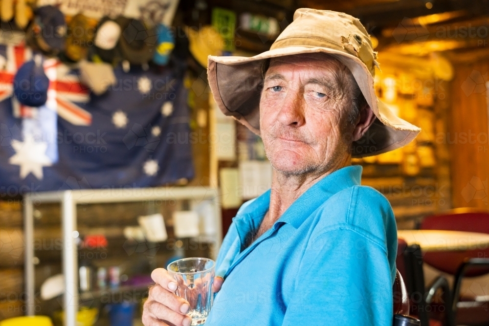 A elderly man sitting in an outback pub holding a glass to his chest - Australian Stock Image