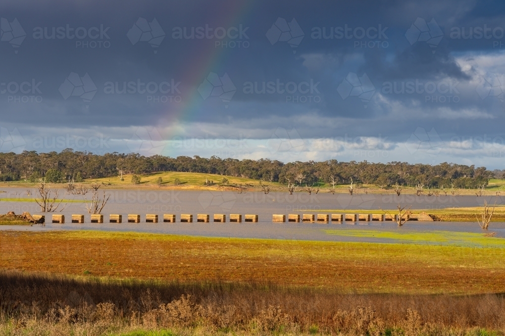 A faint rainbow over a line of concrete pillars rising out of a drying water reservoir - Australian Stock Image