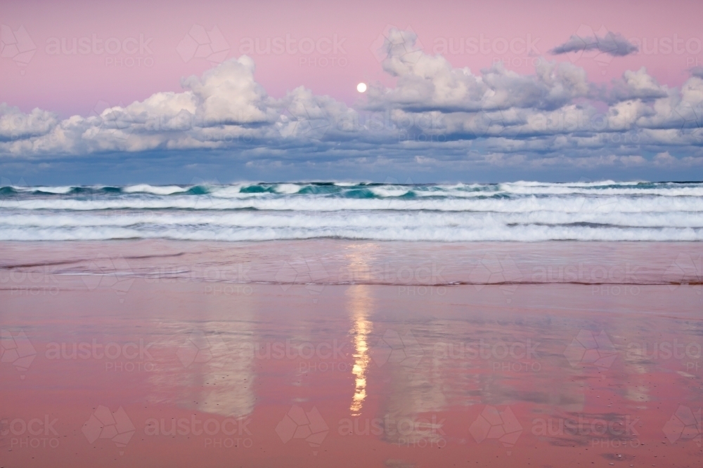 A full Moon rising out of clouds over a beach with colourful reflections on the wet sand - Australian Stock Image