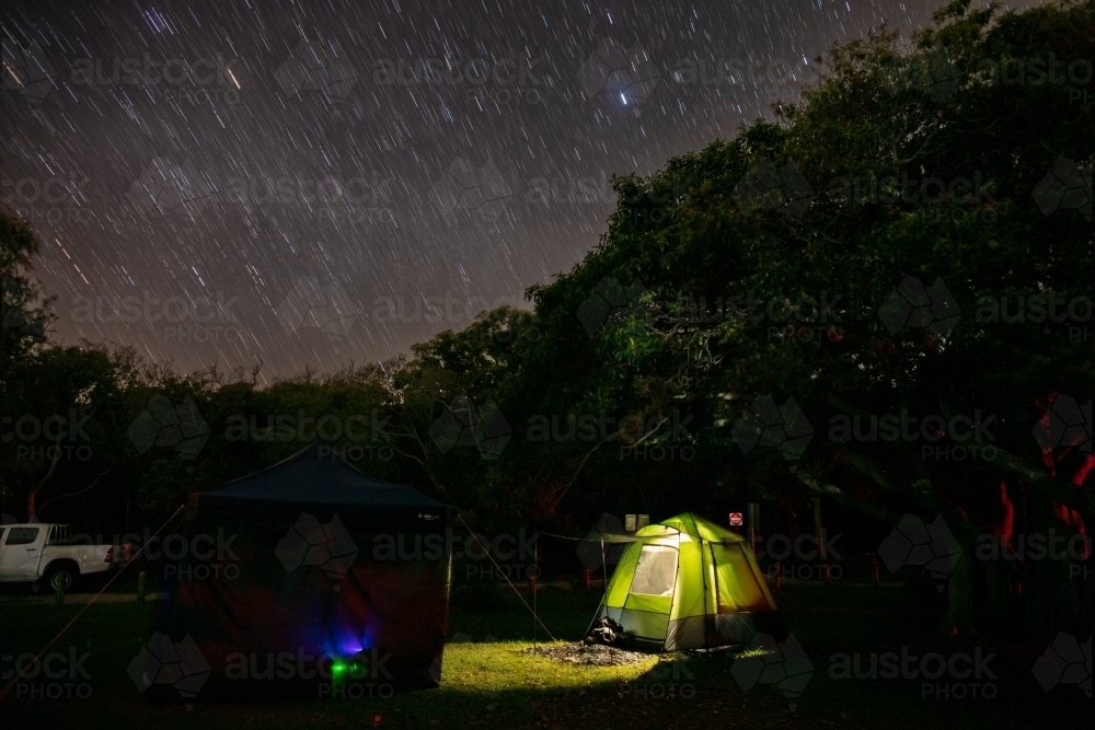 A glowing green tent under a star filled sky (with star trails) and a canopy of dark trees. - Australian Stock Image