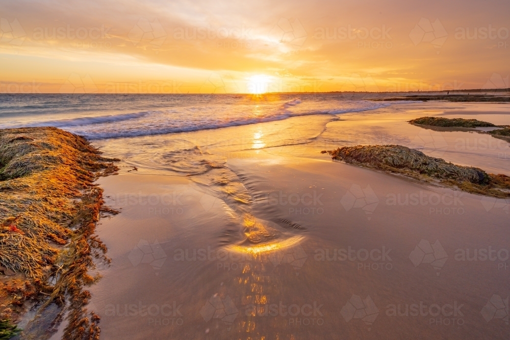 A golden sunset over the ocean reflecting in a wet sandy beach between rocks - Australian Stock Image