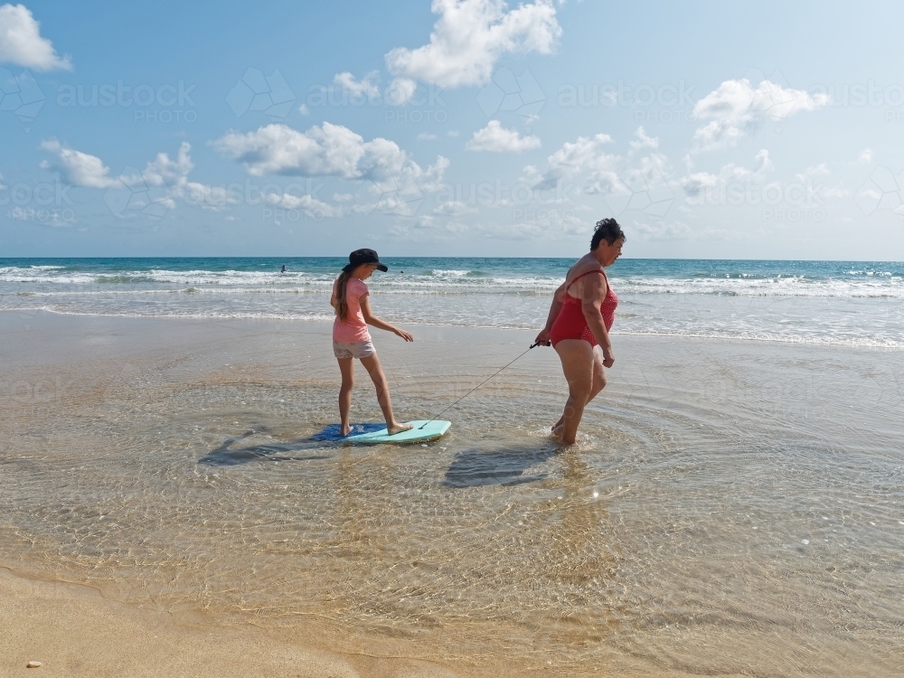A grandmother pulling a girl on a surf board along an empty beach - Australian Stock Image