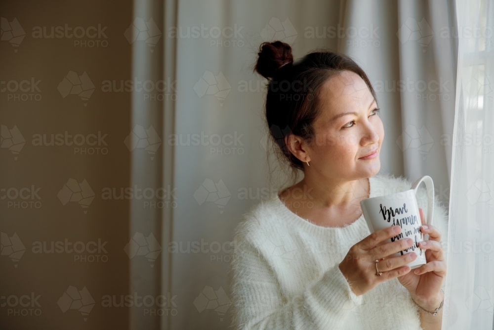 A happy and content woman reflecting next to the window at home - Australian Stock Image