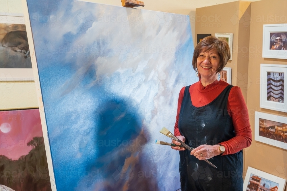 A happy female artist standing in front of a colourful painting with brush in hand - Australian Stock Image